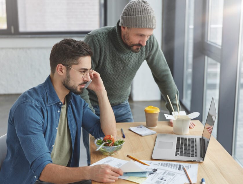 Experienced bearded male and his trainee develop new strategy together, work on improvement business affairs, study information from laptop, ask consultation in colleagues have dinner at office